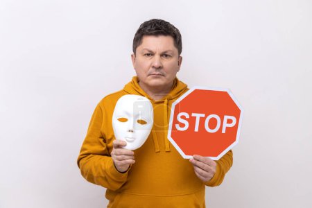 Photo for Portrait of serious dark haired man holding white mask with unknown face and red traffic sign, looking at camera, wearing urban style hoodie. Indoor studio shot isolated on white background. - Royalty Free Image