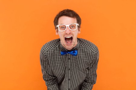 Photo for Emotional scared man nerd looks with frightened expression, looks nervously at camera, demonstrates phobia, wears shirt with blue bow tie and glasses. Indoor studio shot isolated on orange background. - Royalty Free Image