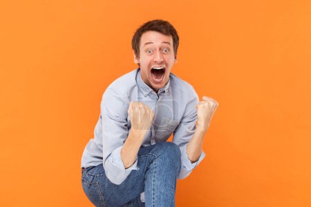 Photo for Portrait of excited amazed young adult man standing screaming happily, clenched fists, yelling with excitement, wearing light blue shirt. Indoor studio shot isolated on orange background. - Royalty Free Image