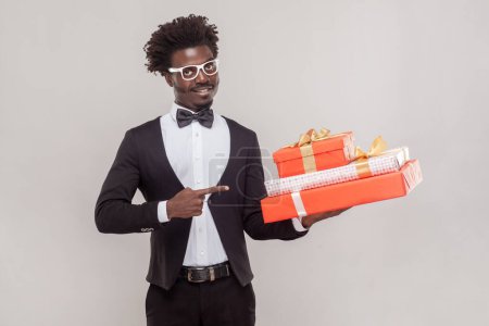 Photo for Portrait of attractive positive young adult man in glasses pointing at three present boxes in his hands, looking at camera, wearing shirt and tuxedo. Indoor studio shot isolated on gray background. - Royalty Free Image