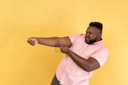 Photo for Portrait of persistent man with beard wearing pink shirt pretending to pull, , holding heavy virtual burden with expression of great effort. Indoor studio shot isolated on yellow background. - Royalty Free Image