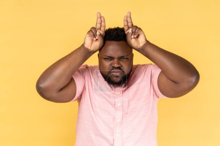 Photo for Portrait of man wearing pink shirt showing bull horn gesture, looking at camera with hostile angry expression, bully male threatening to attack. Indoor studio shot isolated on yellow background. - Royalty Free Image