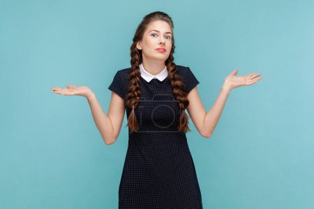Photo for Puzzled uncertain young woman with braids spreads hands aside, shrugging shoulders, don't know how to make a decision, wearing black dress. woman Indoor studio shot isolated on blue background. - Royalty Free Image