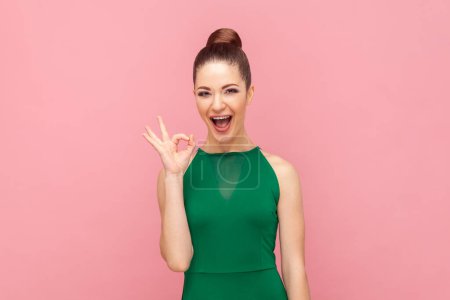 Photo for Portrait of excited amazed young adult woman showing ok sign, looking at camera with positive look, expressing happiness, wearing green dress. Indoor studio shot isolated on pink background. - Royalty Free Image