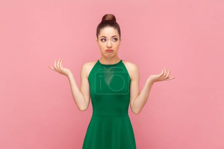 Téléchargez les photos : Portrait d'une femme incertaine confus avec une coiffure de chignon debout avec les mains écartées, regardant la caméra avec une expression incertaine, portant une robe verte. Studio intérieur tourné isolé sur fond rose. - en image libre de droit
