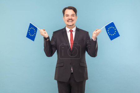 Téléchargez les photos : Portrait d'un homme positif avec une moustache tenant les drapeaux de l'UE et regardant la caméra avec un sourire de dents, portant un costume noir avec une cravate rouge. Studio intérieur tourné isolé sur fond bleu clair. - en image libre de droit
