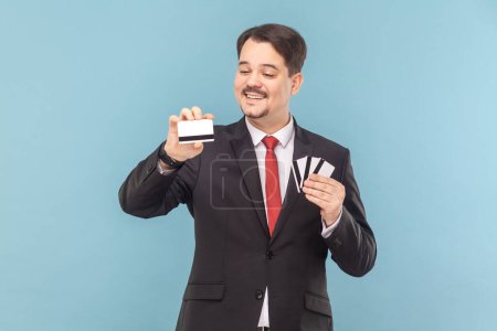 Photo for Portrait of delighted smiling greedy man with mustache standing holding many credit cards with big sum of money, wearing black suit with red tie. Indoor studio shot isolated on light blue background. - Royalty Free Image