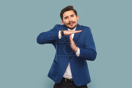 Photo for Portrait of nervous stressed man with mustache standing showing time limit gesture, warning about deadline, wearing official style suit. Indoor studio shot isolated on light blue background. - Royalty Free Image