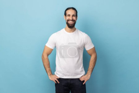 Photo for Portrait of bearded handsome man wearing white T-shirt standing with hands on hips, looking at camera with satisfied face and smiling. Indoor studio shot isolated on blue background. - Royalty Free Image