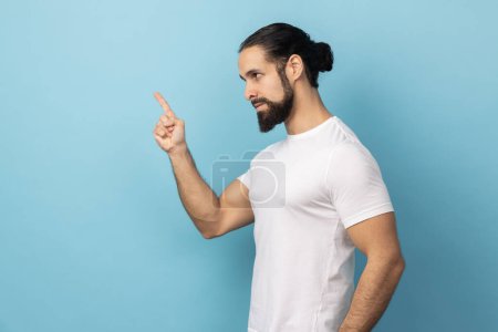 Photo for Side view of angry man with beard wearing T-shirt pointing finger at camera and looking with dissatisfied suspicious expression, warning about troubles. Indoor studio shot isolated on blue background. - Royalty Free Image