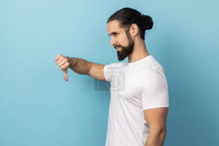 Photo for Side view of extremely dissatisfied man with beard wearing white T-shirt showing thumbs down, dislike and disagree with information, conflict. Indoor studio shot isolated on blue background. - Royalty Free Image