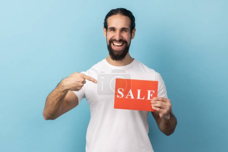 Photo for Look at this. Portrait of extremely happy man with beard wearing white T-shirt pointing finger at sale card in her hand, market discounts. Indoor studio shot isolated on blue background. - Royalty Free Image