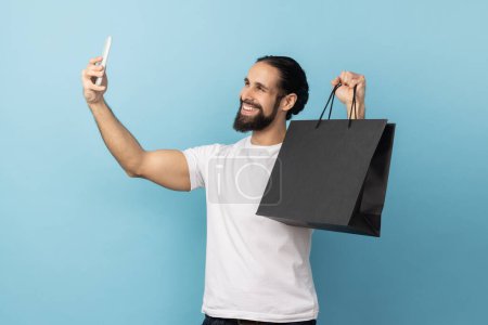 Photo for Portrait of man with beard wearing white T-shirt broadcasting livestream and boasting his purchase, holding shopping, using smartphone. Indoor studio shot isolated on blue background. - Royalty Free Image