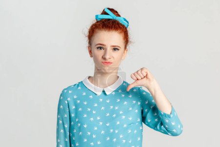 Photo for Portrait of unhappy sad upset red haired woman with hair bun, showing thumb down, being displeased, frowning her face, wearing blue dress. Indoor studio shot isolated on gray background. - Royalty Free Image