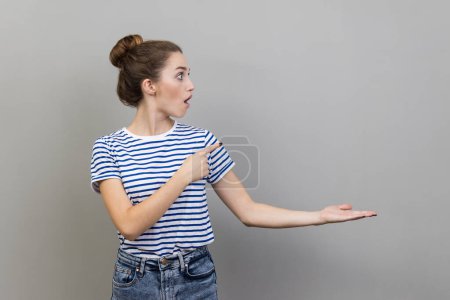 Photo for Wow, product advertisement. Portrait of amazed woman in striped T-shirt presenting copy space on palm, showing empty place for commercial text or goods. Indoor studio shot isolated on gray background. - Royalty Free Image