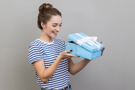 Téléchargez les photos : Portrait de femme portant des rayures T-shirt déballage boîte cadeau bleu, regarder à l'intérieur avec expression faciale positive, cadeau d'anniversaire. Studio intérieur tourné isolé sur fond gris. - en image libre de droit