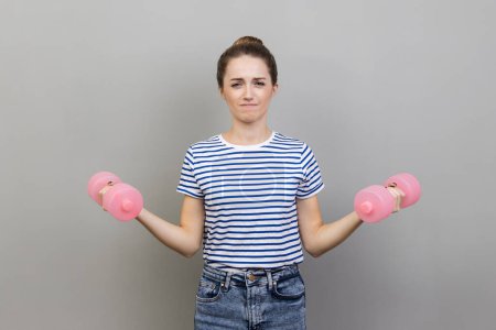 Téléchargez les photos : Portrait de femme portant un T-shirt rayé tenant de petits haltères roses, pompant les muscles, faisant des exercices sportifs avec effort. Studio intérieur tourné isolé sur fond gris. - en image libre de droit