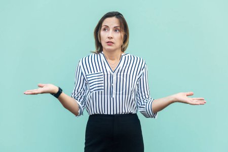 Photo for Portrait of ambiguous confused middle aged woman wearing striped shirt spreads hands aside, being helpless, having uncertain facial expression. Indoor studio shot isolated on light blue background. - Royalty Free Image