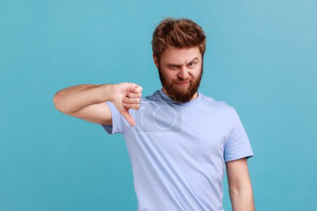 Photo for Portrait of bearded man criticizing bad quality with thumbs down displeased grimace, showing dislike gesture, expressing disapproval. Indoor studio shot isolated on blue background. - Royalty Free Image