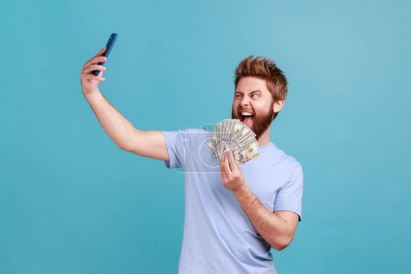 Téléchargez les photos : Portrait d'un bel homme barbu excité faisant du selfie avec un grand fan de billets en dollars, montrant de l'argent aux suiveurs, ayant l'air fou et heureux. Studio intérieur tourné isolé sur fond bleu. - en image libre de droit