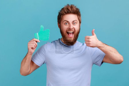 Photo for Portrait of extremely happy optimistic bearded man holding thumbs up paper shape sign and showing like gesture with hand. Indoor studio shot isolated on blue background. - Royalty Free Image