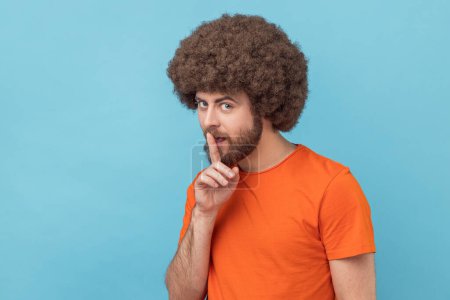 Photo for Portrait of man with Afro hairstyle wearing orange T-shirt showing hush with finger on his lips gesture, shushing, asking to keep silence. Indoor studio shot isolated on blue background. - Royalty Free Image