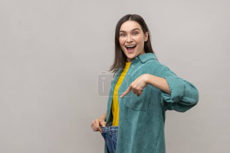 Photo for Beautiful woman have willpower to loose weight, showing slim waist and pointing finger on big trousers, diet conception, wearing casual style jacket. Indoor studio shot isolated on gray background. - Royalty Free Image