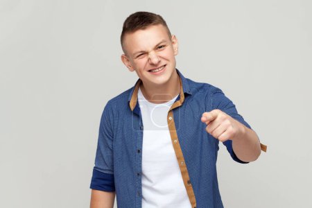 Téléchargez les photos : Portrait d'un adolescent portant une chemise bleue debout et pointant du doigt la caméra, heureux avec le sourire des dents. Studio intérieur tourné isolé sur fond gris. - en image libre de droit