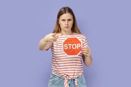 Foto de Retrato de una atractiva mujer rubia seria con una camiseta a rayas sosteniendo una señal de stop rojo y señalando con el dedo a la cámara, mostrando una señal de advertencia con la mano. Estudio interior plano aislado sobre fondo púrpura - Imagen libre de derechos