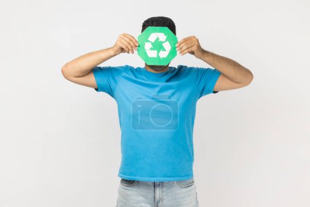 Téléchargez les photos : Portrait d'un homme portant un T-shirt bleu caché derrière un panneau vert de recyclage, de tri des ordures et de protection de l'environnement, pensant vert. Studio intérieur tourné isolé sur fond gris. - en image libre de droit
