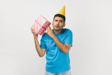 Photo for Portrait of curious unshaven man wearing blue T- shirt and yellow party cone standing shacking present box, being interested what inside. Indoor studio shot isolated on gray background. - Royalty Free Image