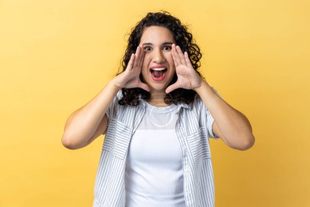Photo for Portrait of winsome young adult woman with dark wavy hair standing and screaming about news or troubles holding her hands near mouth. Indoor studio shot isolated on yellow background. - Royalty Free Image