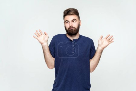 Photo for I Dont Know. Portrait of man with beard wearing dark blue T-shirt looking at camera and having confused facial expression, raised arms. Indoor shot isolated on gray background. - Royalty Free Image