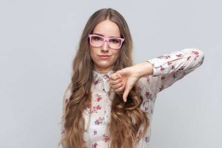 Photo for Portrait of disappointed young adult woman in glasses with long blond hair showing thumb down, expressing bad negative emotions, dislike. Indoor studio shot isolated on gray background. - Royalty Free Image