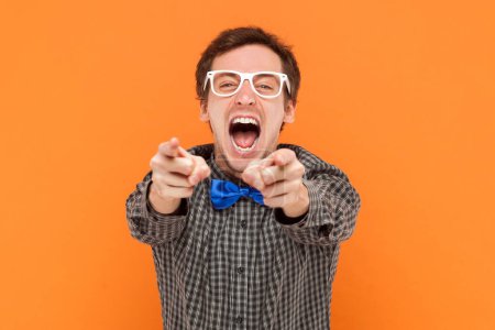 Photo for Portrait of crazy laughing man nerd pointing at you, mocking at you, shame on you, wearing shirt with blue bow tie and white glasses. Indoor studio shot isolated on orange background. - Royalty Free Image