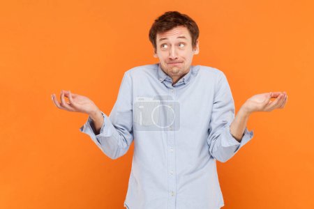 Photo for Portrait of puzzled confused young adult man standing and looikng away, shrugging his shoulders, don't know answer, wearing light blue shirt. Indoor studio shot isolated on orange background. - Royalty Free Image
