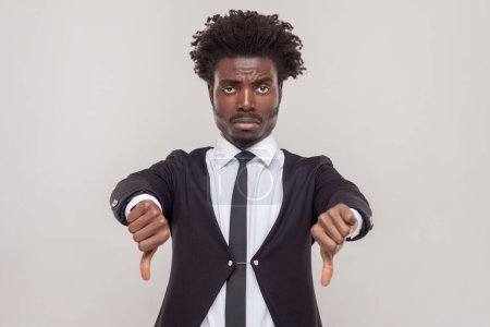 Photo for Man with Afro hairstyle keeps thumb down, dissaproves something, frowns face in discontent, doesn`t like boring film and not recommend to watch it. Indoor studio shot isolated on gray background. - Royalty Free Image