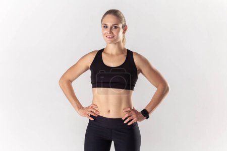 Photo for Pretty beautiful athletic blonde woman standing with hands on hips, looking at camera with positive confident expression, wearing black fitness clothing. Indoor studio shot isolated on gray background - Royalty Free Image