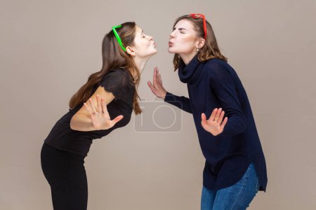 Photo for Side view portrait of two funny happy beautiful women best friends standing with spread hands, sending air kissing to each other. Indoor studio shot isolated on light brown background. - Royalty Free Image