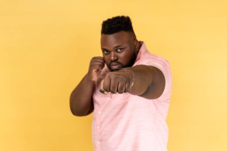Photo for I'll hit you. Portrait of bearded man wearing pink shirt standing with raised fists boxing gesture, ready to punch, ready to struggle, fighting spirit. Indoor studio shot isolated on yellow background - Royalty Free Image