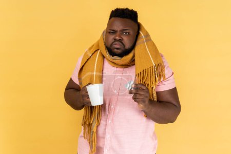 Photo for Portrait of sick unhealthy bearded man wearing pink shirt wrapped in scarf holding tea and pills, feeling despair about influenza. Indoor studio shot isolated on yellow background. - Royalty Free Image