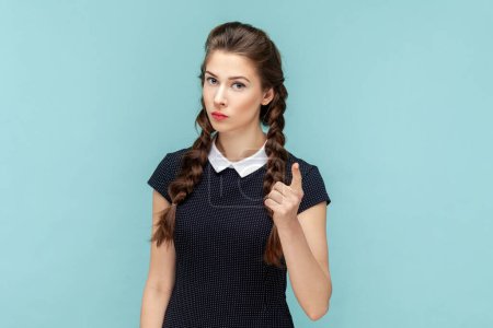 Photo for Portrait of serious strict woman with braids standing and raised index finger, warming you showing disapproval gesture, wearing black dress. woman Indoor studio shot isolated on blue background. - Royalty Free Image