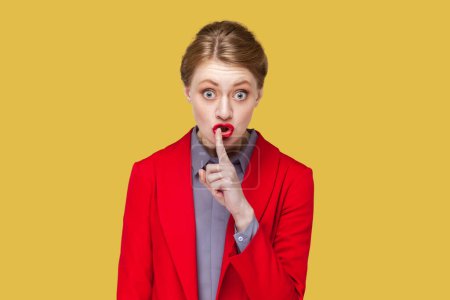 Photo for Portrait of strict bossy woman with red lips standing looking at camera, showing shh gesture, asking to be quiet, wearing red jacket. Indoor studio shot isolated on yellow background. - Royalty Free Image