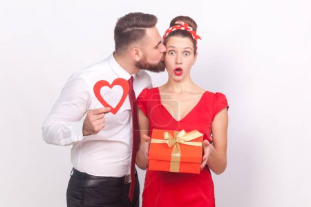 Photo for Shocked woman and romantic man in elegant clothes celebrating anniversary, husband showing red heart, wife looking at camera with amazed face. Indoor studio shot isolated on gray background. - Royalty Free Image