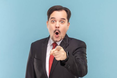 Photo for Shocked surprised astonished man standing and pointing at you, looking at camera with open mouth and big eyes, wearing black suit with red tie. Indoor studio shot isolated on light blue background. - Royalty Free Image