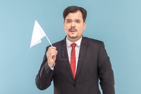 Photo for Portrait of man with mustache standing holding a white flag, looking at camera with scared expression, wearing black suit with red tie. Indoor studio shot isolated on light blue background. - Royalty Free Image