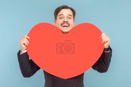 Téléchargez les photos : Portrait d'un homme optimiste extrêmement heureux avec une moustache debout regardant la caméra et tenant un grand cœur rouge, portant un costume noir avec une cravate rouge. Studio intérieur tourné isolé sur fond bleu clair. - en image libre de droit