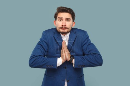 Photo for Portrait of hopeful man with mustache standing with palms together, showing praying gesture, asking apology, wearing white shirt and jacket. Indoor studio shot isolated on light blue background. - Royalty Free Image