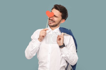 Téléchargez les photos : Portrait d'un bel homme romantique souriant avec une moustache debout couvrant son œil avec un petit cœur rouge sur le bâton, portant une photo de studio intérieure isolée sur fond bleu clair. - en image libre de droit
