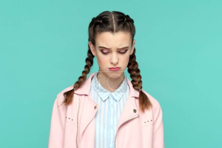 Photo for Portrait of sad upset teenager girl with braids wearing pink jacket standing with head down, expressing sorrow ans sadness, being in bad mood. Indoor studio shot isolated on green background. - Royalty Free Image